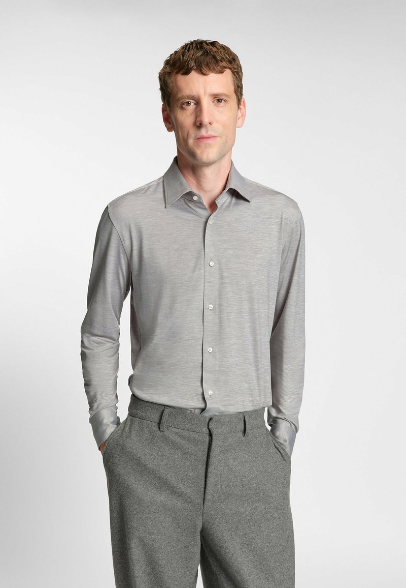 Camicia grigia a maniche lunghe, con texture liscia, colletto classico e apertura con bottoni; abbinata a pantaloni grigi con tasche laterali.