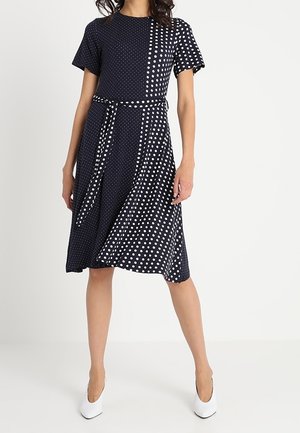 Femme portant une robe bleu marine mi-longue à pois blancs de tailles petites et grandes mélangées, manches courtes, ceinture à la taille, et des talons blancs.