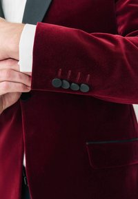 Burgundi bársony zakó fekete hajtókával, hat fekete szövet gombbal az ujjan és egy elegáns zseb részlettel.