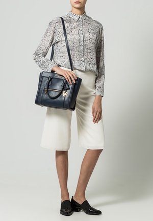 Femme en blouse à manches longues à motifs, short blanc mi-cuisse, mocassins noirs, portant un sac à main en cuir noir avec des accents dorés.