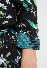 Svart tygsleeve med elastisk mudd som har gröna och vita blommor och bladmönster på en persons arm mot en enfärgad bakgrund.