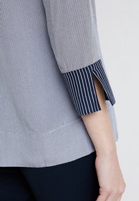 Bluzka w paski w jasno- i ciemnoniebieskim kolorze, z rękawami o długości trzy czwarte z rozcięciami oraz gładką teksturą materiału. Brzeg jest nieco dłuższy z tyłu.