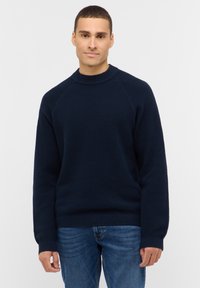 Dunkelblauer Strickpullover mit strukturiertem Muster, rundem Ausschnitt und gerippten Bündchen und Saum. Getragen mit blauen Jeans.