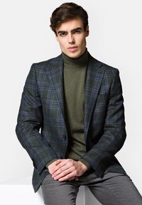 Marynarka w ciemnozieloną kratę z niebieskimi akcentami, z klapami i dwoma guzikami. Nosi ją na dopasowanym oliwkowym golfie. Szare spodnie.