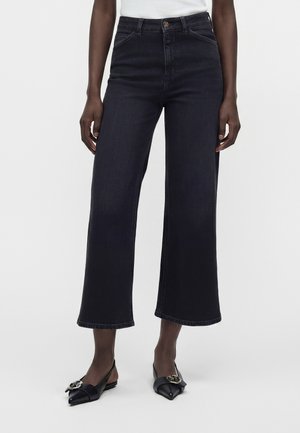 Person iført mørke, vide cropped jeans og sorte spidse slingback-sko med gulddetaljer på spænde på en hvid baggrund.