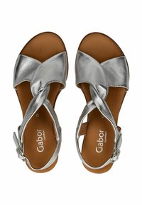 Sandalias de cuero plateado con un diseño superior retorcido, plantilla acolchada marrón y correa en el tobillo ajustable con una hebilla plateada.
