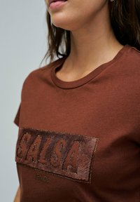 Camiseta de algodón marrón con escote redondo y mangas cortas, con un logotipo texturizado "SALSA" en marrón más oscuro y acentos de tachuelas.