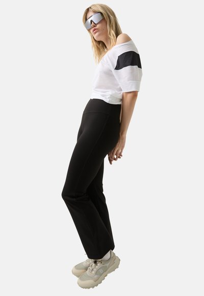 Witte off-shoulder top met zwarte streep, zwarte, aansluitende broek en beige sportieve schoenen. Model draagt een oversized zilveren zonnebril.