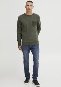 Oliven grønn hettegenser med en teksturert lomme, kombinert med blå jeans og svarte joggesko. Designet har rund hals og ribbestrikkede ermer.