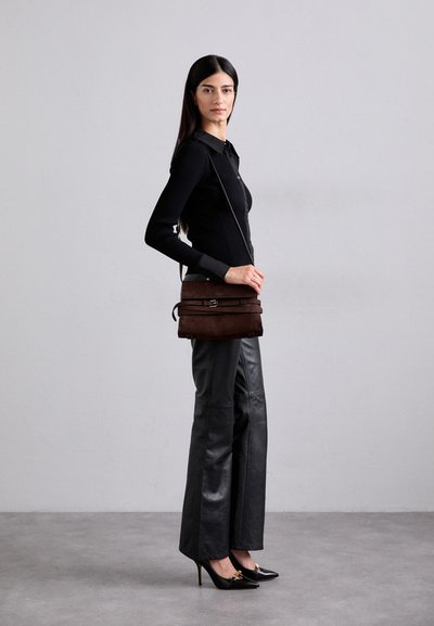 Bolso de ante marrón con un diseño plano, que presenta un detalle de hebilla, llevado en bandolera. Modelo con top de canalé negro y pantalones de cuero.