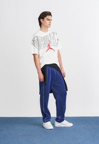 Valkoinen lyhythihainen t-paita punaisella Jordan-logolla, yhdistettynä sinisiin fleece-housuihin, joissa on sivuviivat, sekä valkoisiin tennareihin.