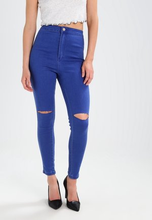 Frau trägt high-waisted blaue zerrissene Skinny-Jeans, weißes bauchfreies Top und schwarze High-Heels, steht vor einem schlichten Hintergrund.