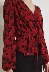 Blouse à motifs floraux rouges avec manches longues, décolleté en V et taille froncée nouée avec un ruban. Tissu doux avec une silhouette fluide.