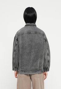 Pessoa com cabelo preto liso, vestindo uma jaqueta de ganga preta oversized e desbotada, e calças bege, de costas contra um fundo branco liso.