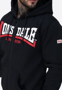 Sudadera con cremallera negra hecha de algodón, con un logo de Lonsdale en rojo y blanco en el pecho y un pequeño parche de logo en la manga.