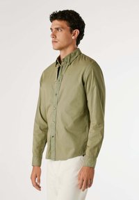 Camicia verde oliva a maniche lunghe, realizzata in cotone leggero, presenta un colletto classico, bottoni a vista e pantaloni bianchi.