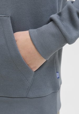 Roka delno v žepu modro-sivega puloverja z rebrastimi manšeti in pasom, ki prikazuje teksturo blaga in podrobnosti šivanja.