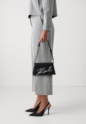 Schwarze Handtasche mit einer Kettengriff und einem Strass-„Karl“-Logo, kombiniert mit einem schwarz-weißen Hahnentrittmuster-Anzug und spitzen, schwarzen Absätzen.