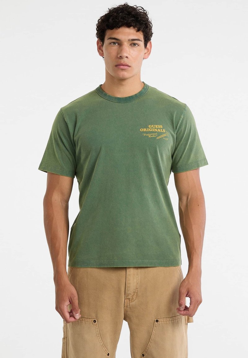 Grünes Baumwoll-T-Shirt mit Rundhalsausschnitt, kurzen Ärmeln und gelbem Logo-Text auf der Vorderseite. Die Textur wirkt weich und hat einen leicht abgetragenen Look.