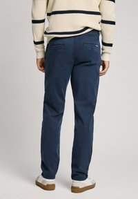 Tamsiai mėlynos medvilnės kelnės su laisvu pjaustymu, turinčios dvi galines kišenes, dėvimos su smėlio spalvos megztiniu ir baltais sportbačiais.