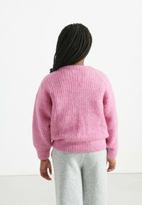 Ružový pletený sveter s rebrovanou textúrou, okrúhlym výstrihom a mierne balónovými rukávmi, v kombinácii so šedými teplákmi. Tkanina vyzerá mäkko a nadýchane.