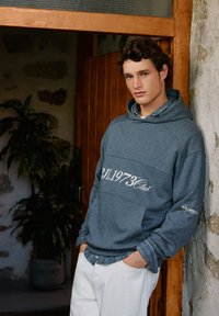 Siva kapuca z logo, z sprednjim žepom in pritrjeno kapuco, plasten preko modre kockaste srajce, v kombinaciji z belimi hlačami.