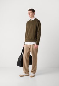 Sweatshirts vert olive, pantalons beiges et baskets blanches. Présente un logo, tient un sac noir, affiché sur un fond neutre.