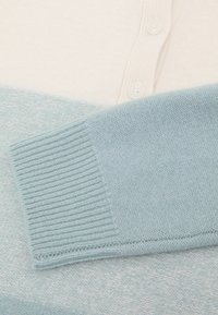 Ľahký pulóverový sveter s krémovou hornou časťou a svetlomodrou spodnou časťou. Má rebrované manžety, gombíkový pás a jemnú textúru.