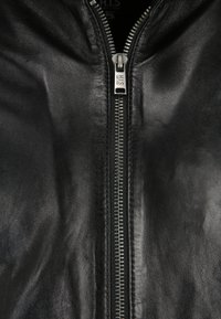 Zwarte leren jas met een rits aan de voorkant, met een gladde textuur en zilverkleurige hardware. Label zichtbaar bij het nekgebied.