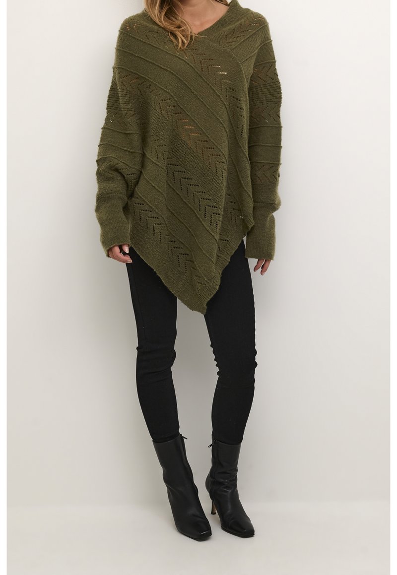 Poncho en tricot de couleur vert olive avec un col en V, une texture géométrique et des rayures diagonales. Assorti à un jean skinny noir et des bottines.