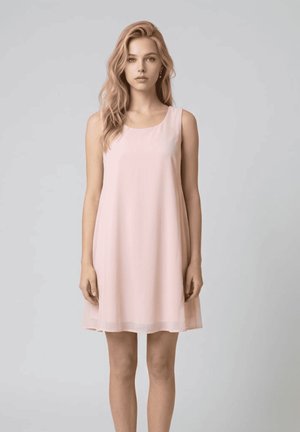 Joven mujer con cabello rubio largo y ondulado que lleva un vestido sin mangas de color rosa claro hasta la rodilla, de pie frente a un fondo gris liso.