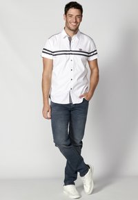Camisa blanca de botones de manga corta con rayas negras, combinada con jeans oscuros y zapatillas blancas. La camisa tiene un logo en el pecho y botones.