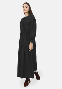 Robe noire à manches longues avec fermeture éclair, deux poches, taille cintrée et design de jupe à volants. Fabriquée dans un tissu lisse.