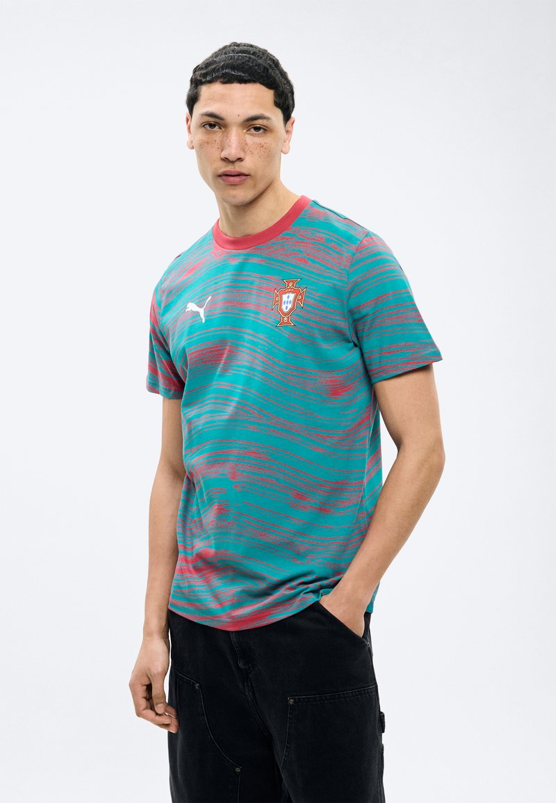 Mees, kes kannab türkiissinist ja punast laineline triibulist Portugal jalgpallimängija särki Puma logoga, seisab ühe käe mustade pükste taskus tavalise tausta ees.