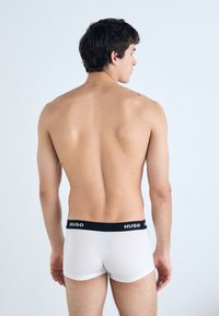 Weiße Baumwoll-Boxershorts mit einem schwarzen Bund, auf dem das Branding "HUGO" zu sehen ist, von hinten vor einem hellen Hintergrund betrachtet.