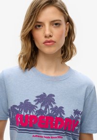 Jasnoniebieska bawełniana koszulka z grafiką palm w ciemnopurpurowym kolorze, z wyrazistym różowym napisem "SUPERDRY". Klasyczny fason z okrągłym dekoltem.