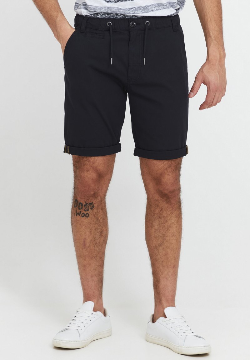 Marineblå shorts laget av lett materiale, med strammelås i livet, rullet kant og sidelommer. Brukt med hvite joggesko.