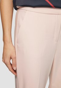 Pantalons sur mesure rose clair avec une texture lisse, poches latérales et un design épuré. Présentés avec une main reposant sur la taille.