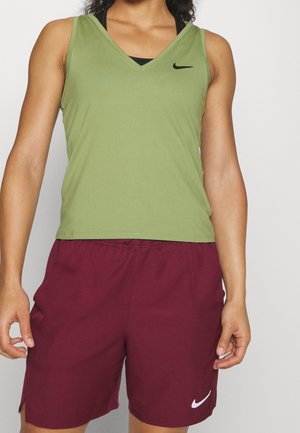 Athlète portant un débardeur vert olive avec un logo noir et un short de sport bordeaux avec un logo blanc, debout devant un fond uni.