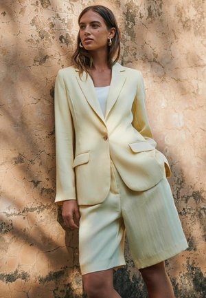 Žena má na sebe svetložlté sako a zodpovedajúce krátke nohavice do kolien, stojí pri štruktúrovanej béžovej stene s rukami v vreckách.