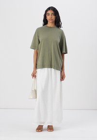 Olijfgroen oversized t-shirt met korte mouwen, gecombineerd met een witte kanten maxi rok. Het model houdt een kleine witte handtas vast en draagt tan sandalen.