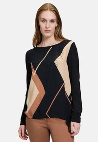 Langärmliges Oberteil mit schwarzen, sandfarbenen und braunen geometrischen Mustern, verziert und mit einem Kordelzug am Saum. Glatte Stofftextur.