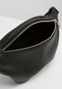 Marsupio in pelle nera con una texture liscia e leggermente granulosa, dotato di una zip argentata e di una tasca interna. Design curvo e tracolla regolabile.