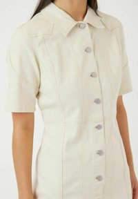 Robe courte beige avec un col, des manches courtes et cinq boutons sur le devant. Présente des coutures verticales et une silhouette ajustée.