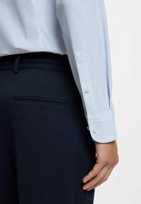 Vaaleansininen ruudullinen paita, jossa on napilliset kalvosimet, yhdistetty tummansinisiin teksturoituihin housuihin, joissa on sivutasku. Siisti ja räätälöity ilme.