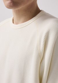 Off-white textured sweatshirt with a crew neck and raglan sleeves, featuring a waffle knit pattern and contrasting stitch details.