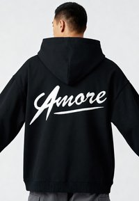 Felpa nera con cappuccio, caratterizzata da una prominente grafica bianca in corsivo "Amore" sulla schiena. Il tessuto sembra morbido, con polsini e orlo a costine.