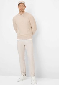 Mann trägt beigen Pullover, passende Hose, weiße Turnschuhe und beige Mütze, steht vor einem schlichten weißen Hintergrund mit den Händen hinter dem Rücken.