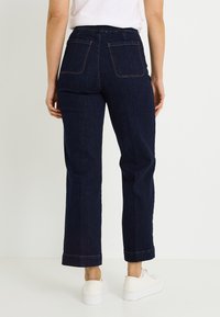 Pantalones vaqueros de color azul oscuro con un diseño de pierna recta, cintura alta, costuras naranjas y dos bolsillos traseros. Calzado: zapatillas blancas.