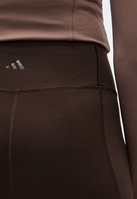 Gros plan d'une personne portant un legging de sport taille haute marron foncé avec un petit logo réfléchissant Adidas sur la ceinture.